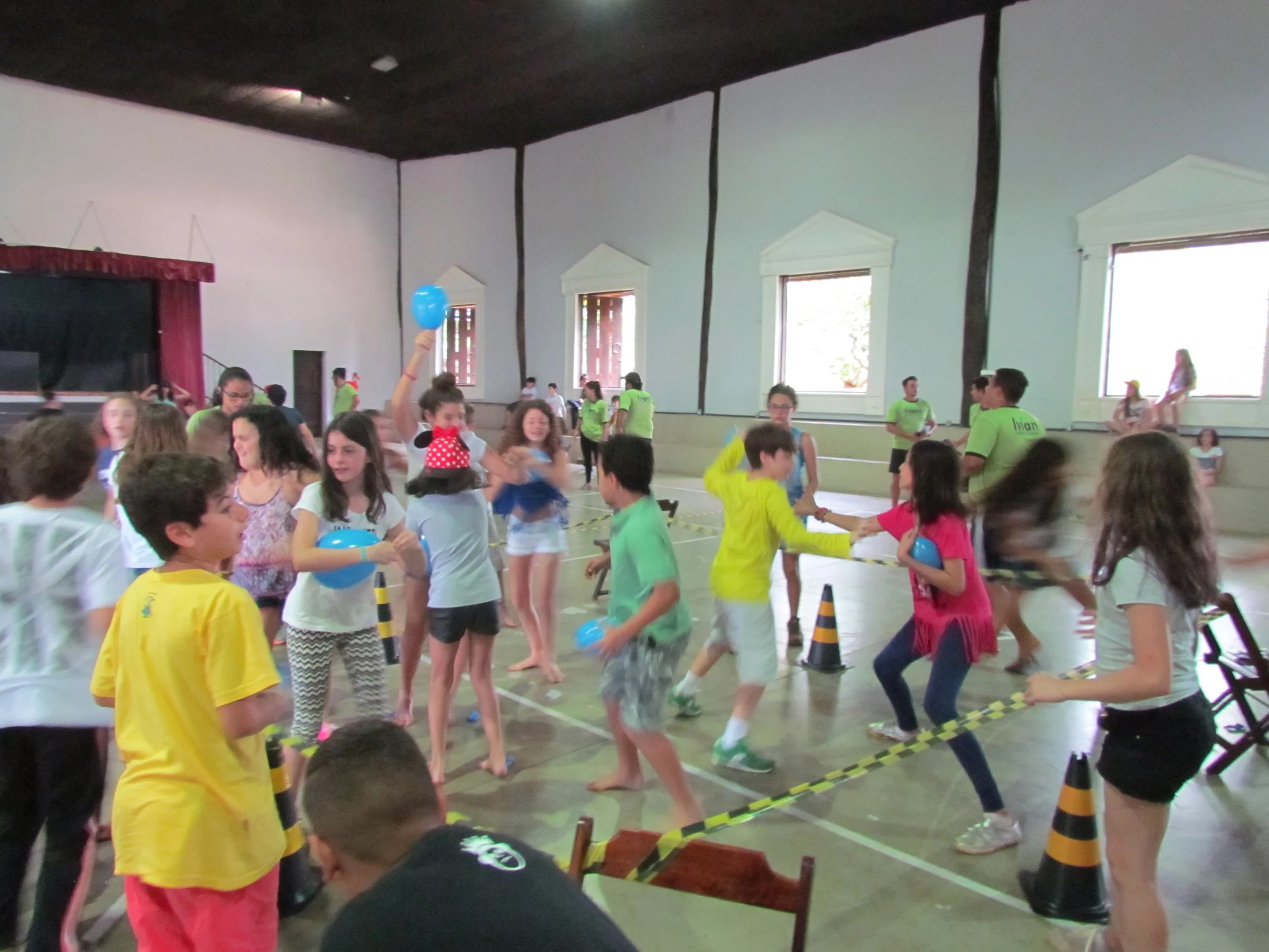 Crianças brincando em um ginásio coberto, com algumas segurando balões azuis e vestidas com roupas coloridas, enquanto adultos supervisionam a atividade.