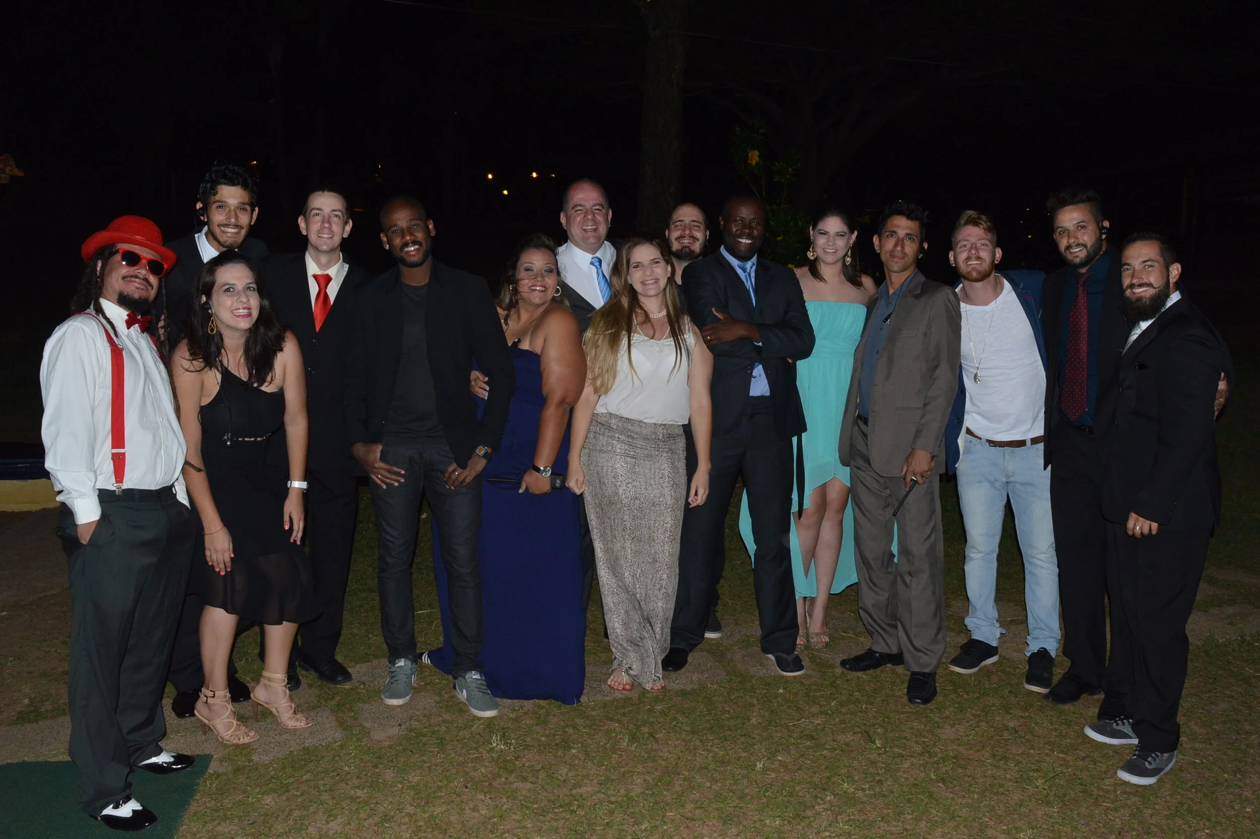 Grupo de pessoas posando para uma foto em um evento noturno ao ar livre, com roupas formais e sorrindo.