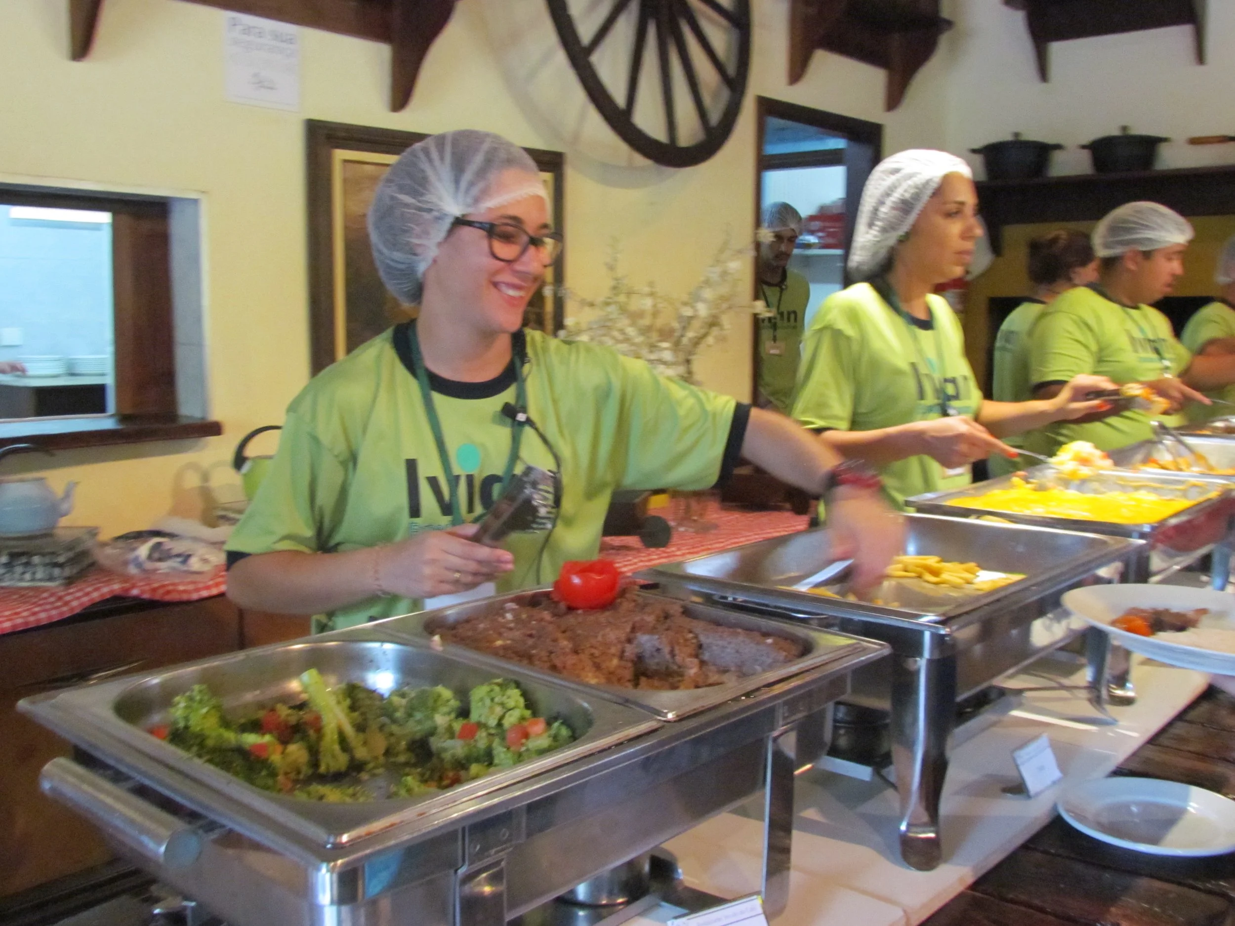 Equipe de pessoas usando uniformes verdes, toucas de cabelo e aventais servindo comida em um bufê. Pratos com salada e alimentos variados na frente.