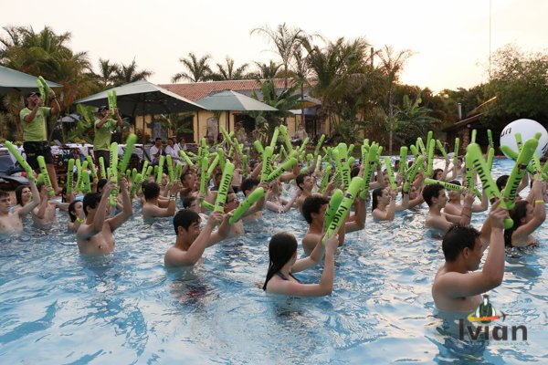 Grupo de crianças em uma piscina ao pôr do sol, brincando com palmas infláveis verdes, com adultos ao fundo e árvores ao redor.