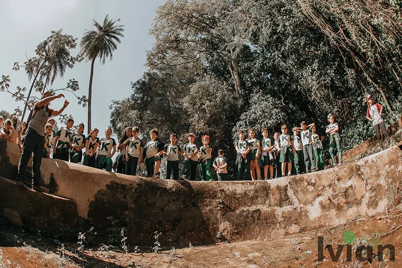 Grupo de estudantes em uniforme escolar, de pé em uma parede de concreto ao ar livre, sob árvores, durante o dia.