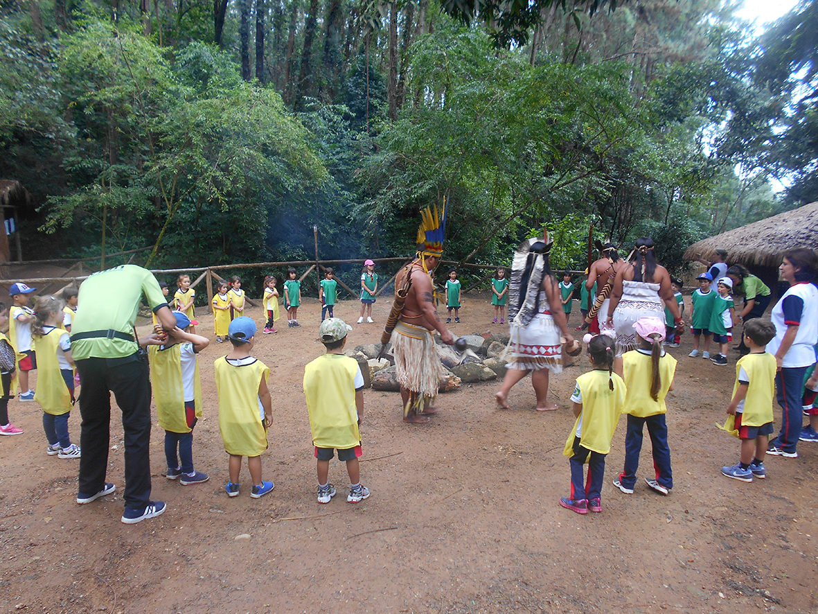 Grupo de crianças participando de uma atividade cultural indígena em um espaço ao ar livre com árvores ao fundo, rodeadas por adultos e indígenas com trajes tradicionais ao redor de uma fogueira.
