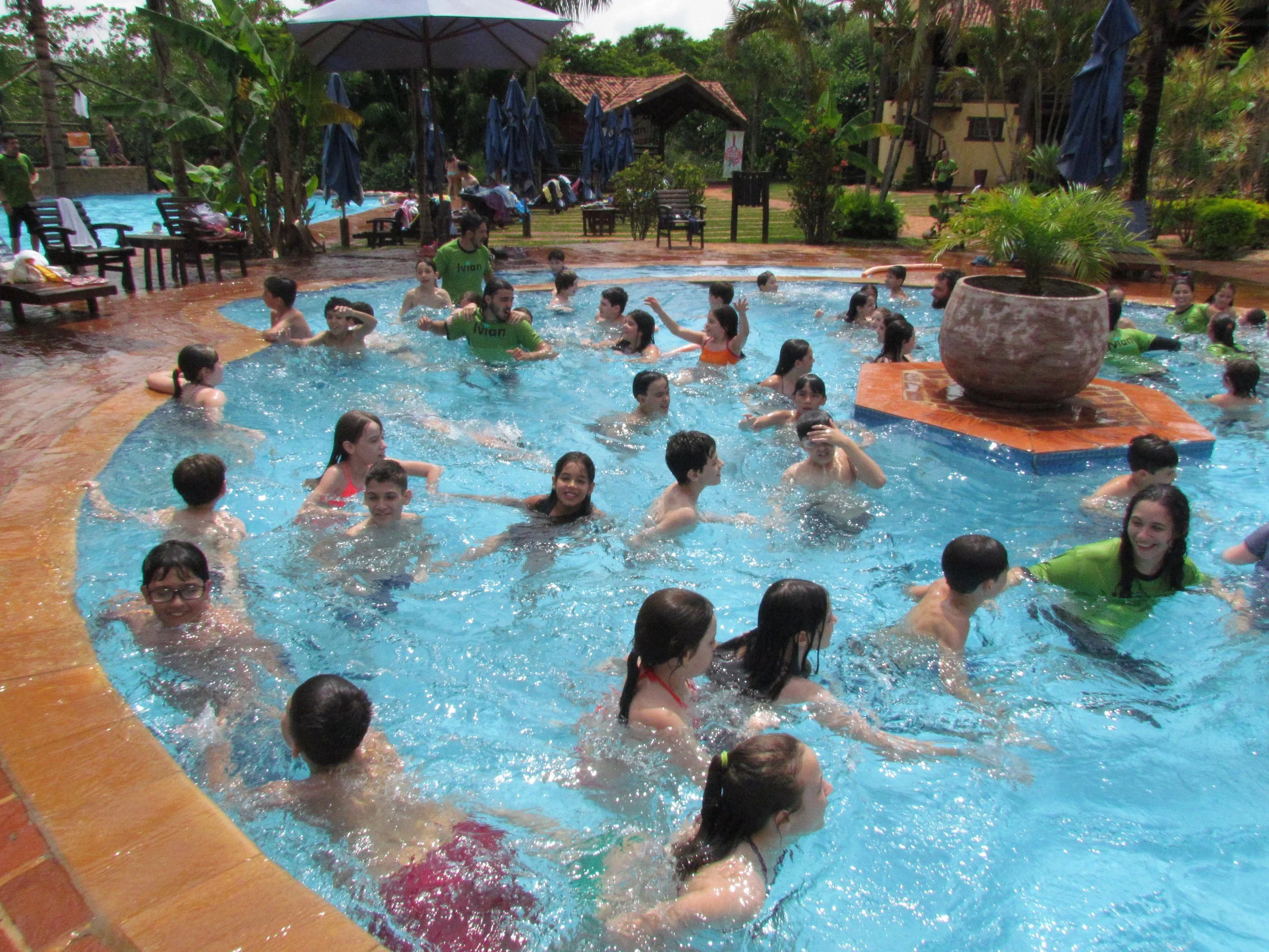 Pessoas brincando e nadando em uma piscina ao ar livre, com árvores e plantas ao redor, brinquedos e espreguiçadeiras nas laterais.