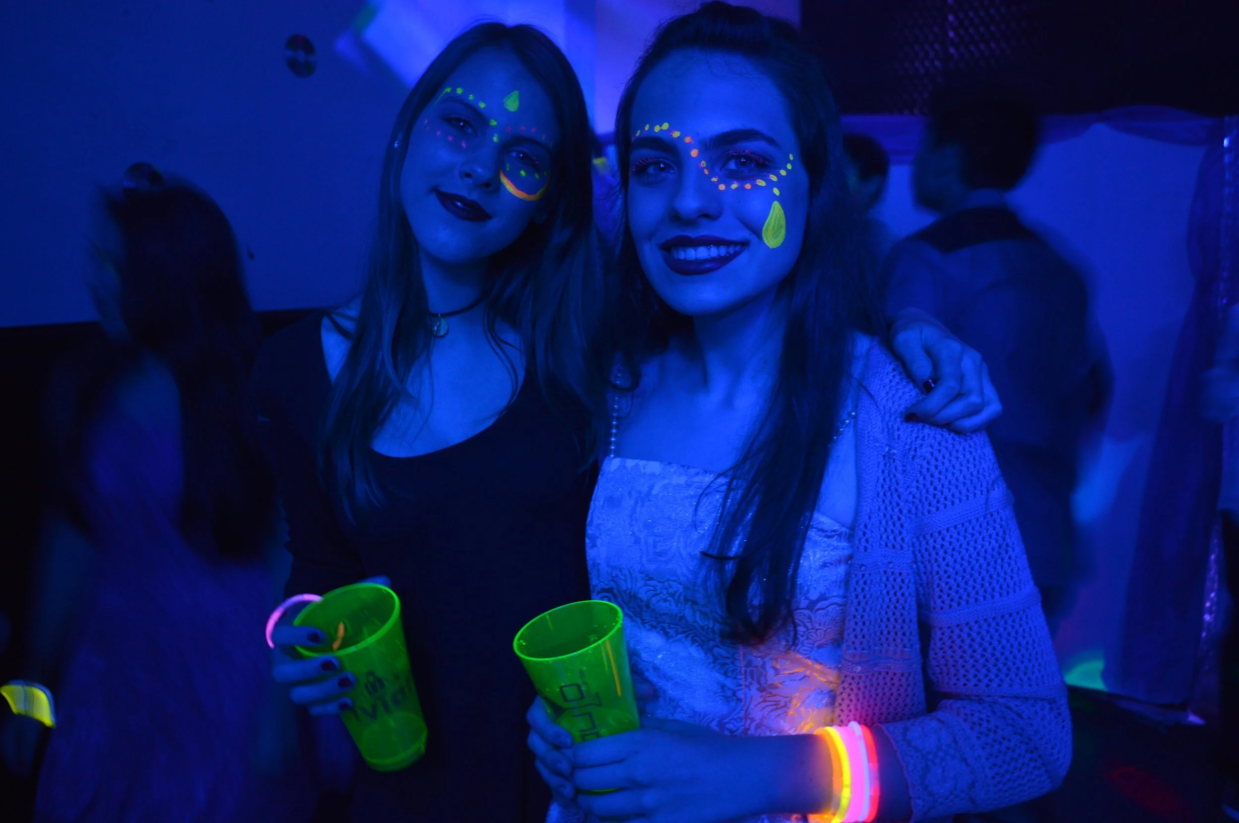 Duas mulheres com maquiagem fluorescente e acessórios luminosos posando em ambiente com luz negra, possivelmente em uma festa ou evento noturno.