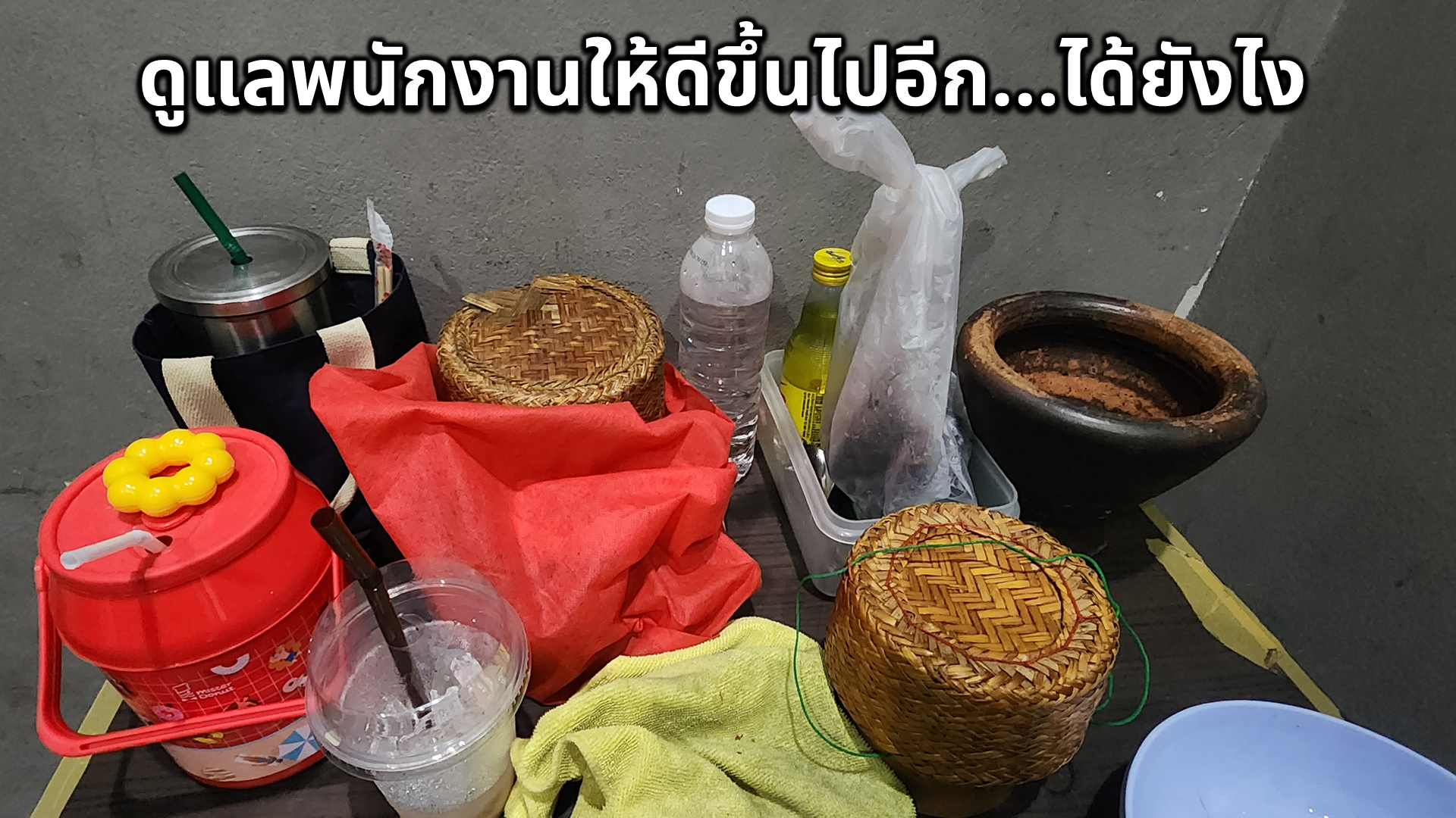 A table holding various items including a water bottle, a yellow container, a plastic bag, two woven baskets, a black container with a straw, a red container with a yellow flower-shaped lid, a clear plastic cup with a straw, and a yellow cloth, with a dark wall in the background.