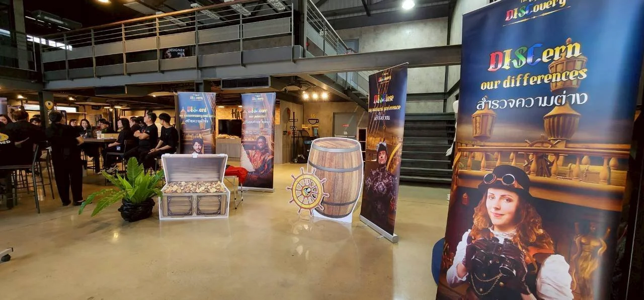 An indoor event space with people seated at a long table on the left and standing around. There are promotional banners with pirate-themed images and the words 'DISCern our differences' and ’สร้างความแตกต่าง’ in Thai, along with a large prop of a barrel, a treasure chest, and a ship’s steering wheel.