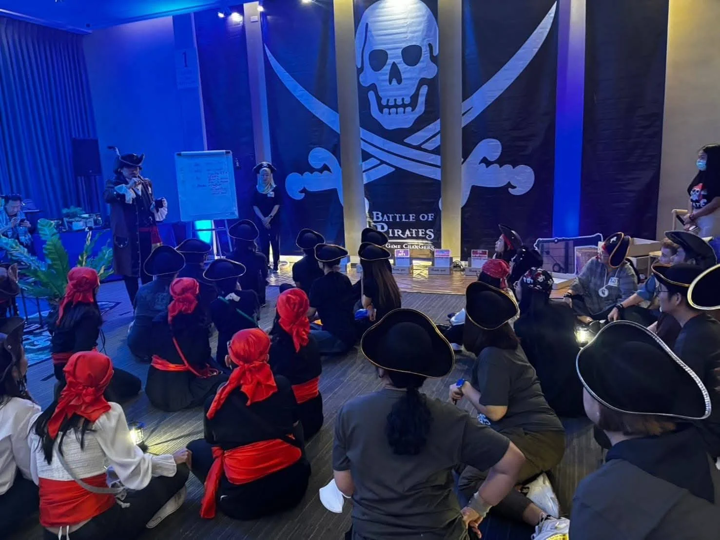 Group of people dressed as pirates attending a themed event with a large pirate flag and skull and crossbones backdrop.