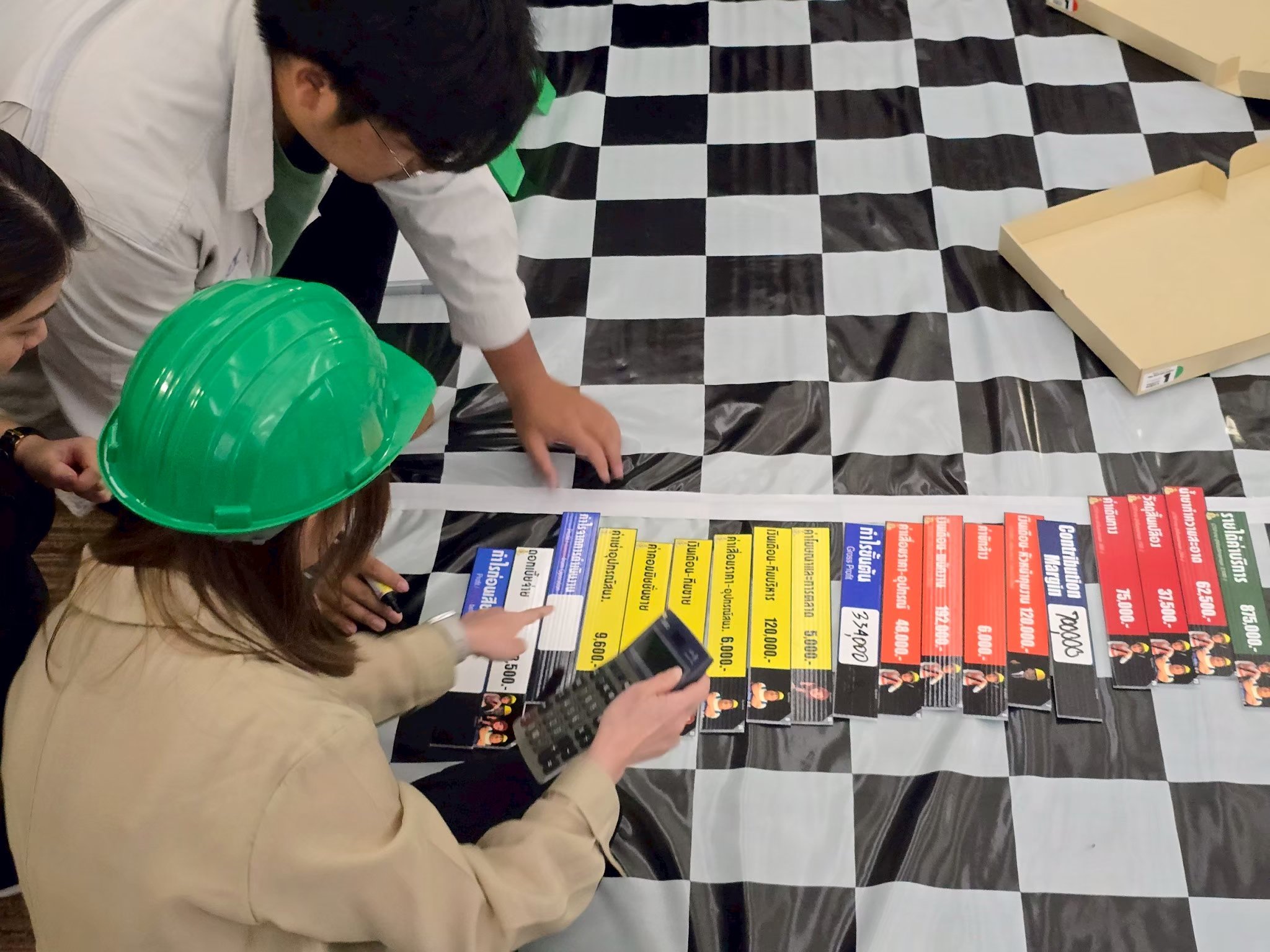 People working together on a project with colorful name tags and a calculator, on a black and white checkered tablecloth, with some empty cardboard boxes in the background.