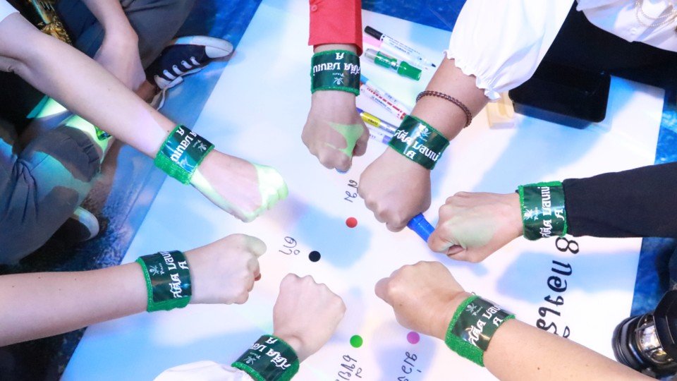 Six people with green wristbands on their wrists, gathered around a table, each with a clenched fist pointing toward the center with colored dots on the table. Some of their hands have paint or marker on them.