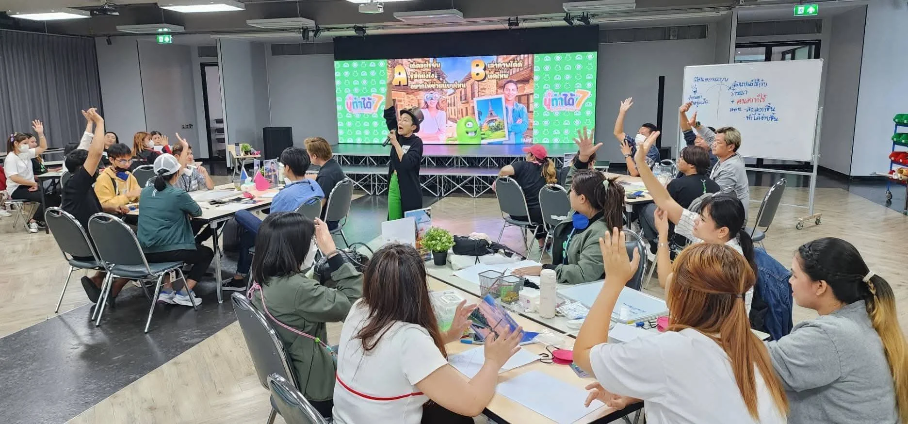 A group of people attending a workshop or conference in a large room with round tables and chairs, raising their hands to ask questions or participate, with a presenter standing at the front near a large digital screen displaying colorful graphics and text, and a whiteboard on the side.