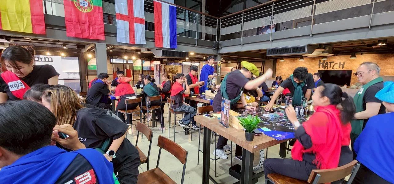 People gathered in an indoor space for a game night, playing board games at tables. Flags of different countries are hanging from the ceiling. The space has a modern industrial design with brick walls and metal railings.