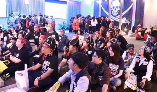 People attending an indoor event with pirate-themed decorations, including a large skull banner with crossbones, and a group of people sitting at tables and others standing in background.