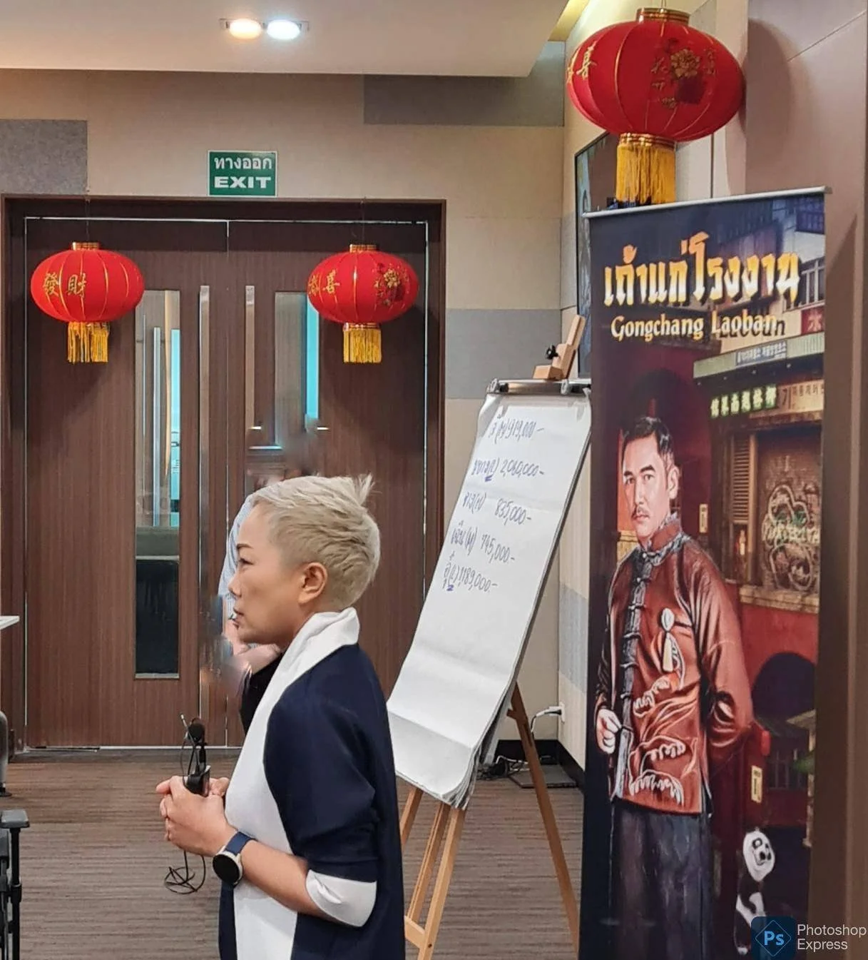 A woman with short blonde hair wearing a navy blazer and white shirt holds a pair of sunglasses in her hand. Behind her are red Chinese lanterns hanging from the ceiling and a large promotional poster featuring a man dressed in traditional Asian clothing. There is also an easel with a whiteboard and a doorway labeled "EXIT" in both Thai and English.