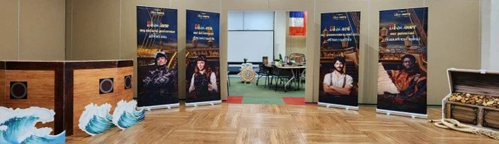An indoor room with a wooden floor, decorated with banners featuring people and scenery, and a small table with chairs in the background.