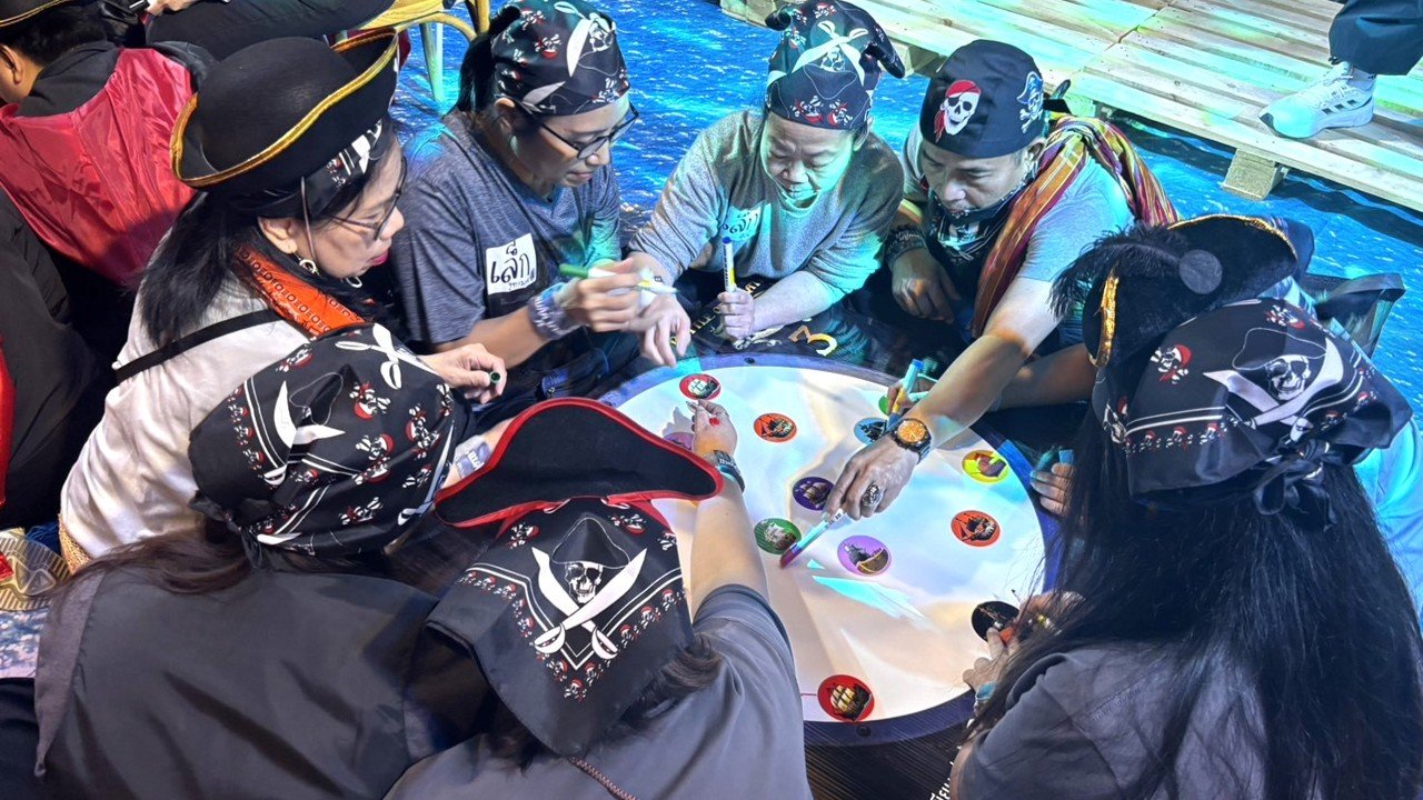 Group of people dressed in pirate costumes playing a board game on a round table with a digital interface, surrounded by water and wooden planks.