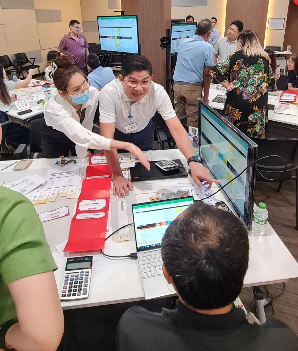 Group of people working and discussing at a busy office or workshop with computers, monitors, and paperwork.