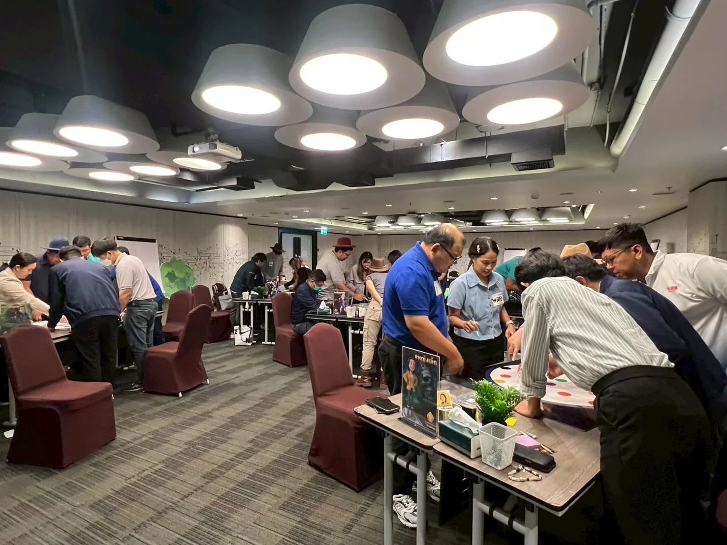 A group of people gathered around tables in a conference room, playing a game on a large, colorful board with dots. Some attendees are standing, leaning over, and engaging in the activity, while others sit observing. The room has modern lighting fixtures on the ceiling and a decorated wall in the background.