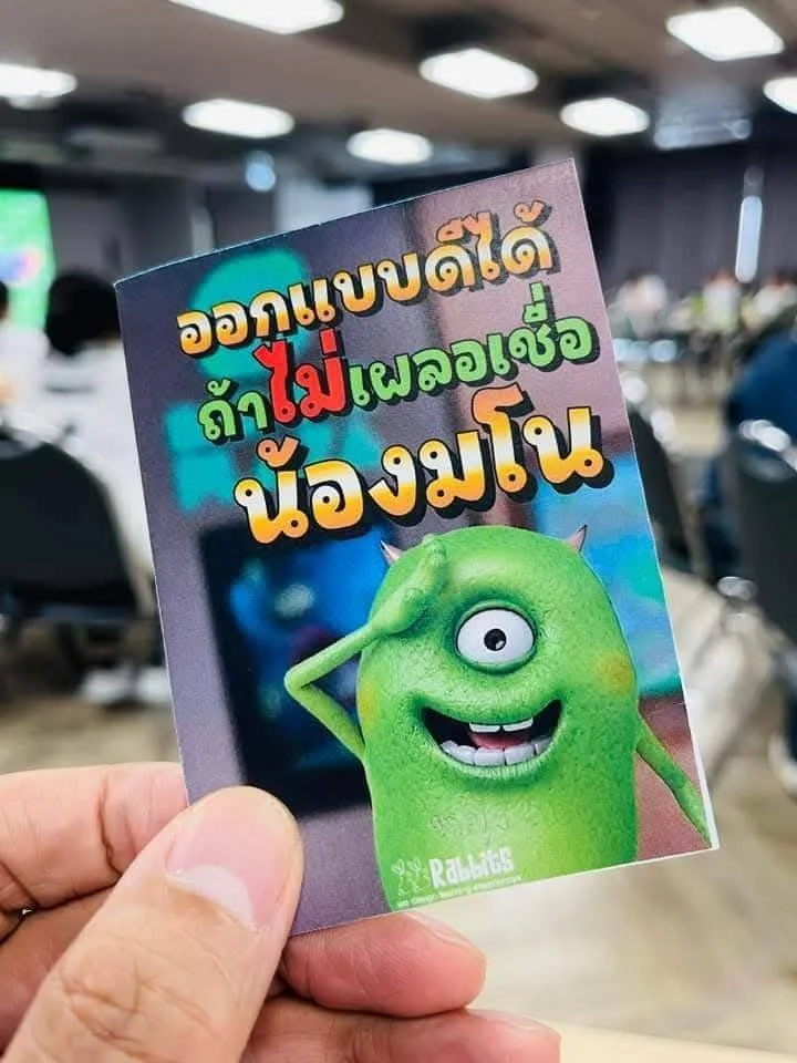 A hand holding a colorful card with Thai text and a cartoon green monster character. The background shows a blurred conference or event room with seated attendees.