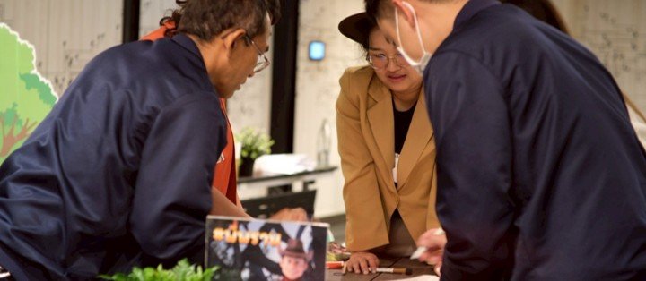 Three people gathered closely at a table, engaged in a discussion or activity, with a map or poster on the wall behind them.