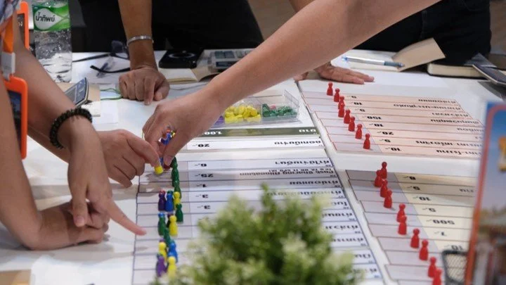 Finance for non-finance course, class vibes-6 | People playing a strategic board game involving colored pieces on a table with score sheets, markers, and a small plant in the foreground.