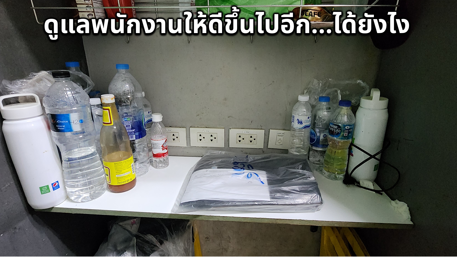 A cluttered countertop with several water bottles, a plastic bag covering some items, electrical outlets on the wall, and chargers or cords, with Thai text overlay at the top.