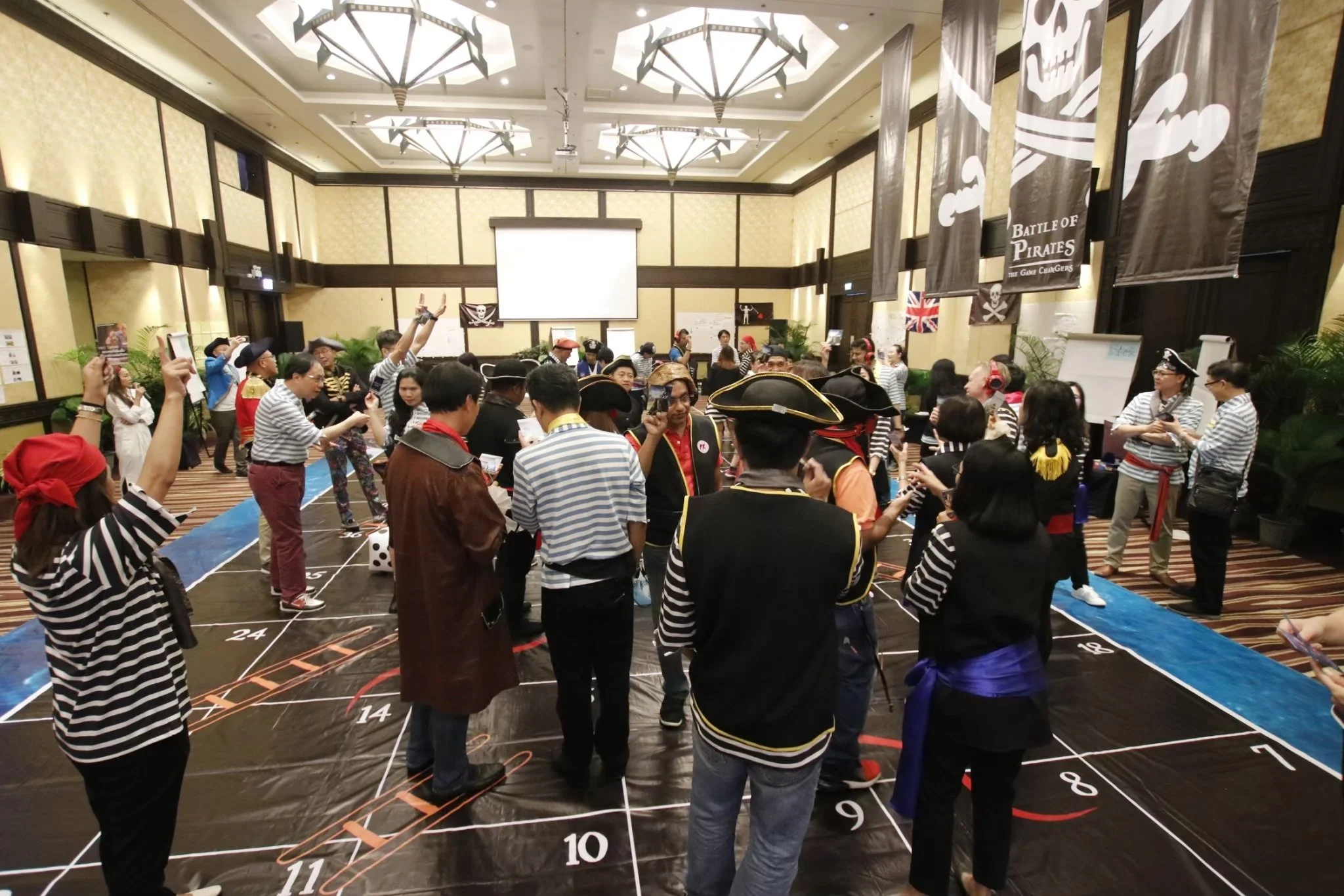 People in a large conference room dressed as pirates, participating in a themed event with pirate banners and a game setup on the floor.