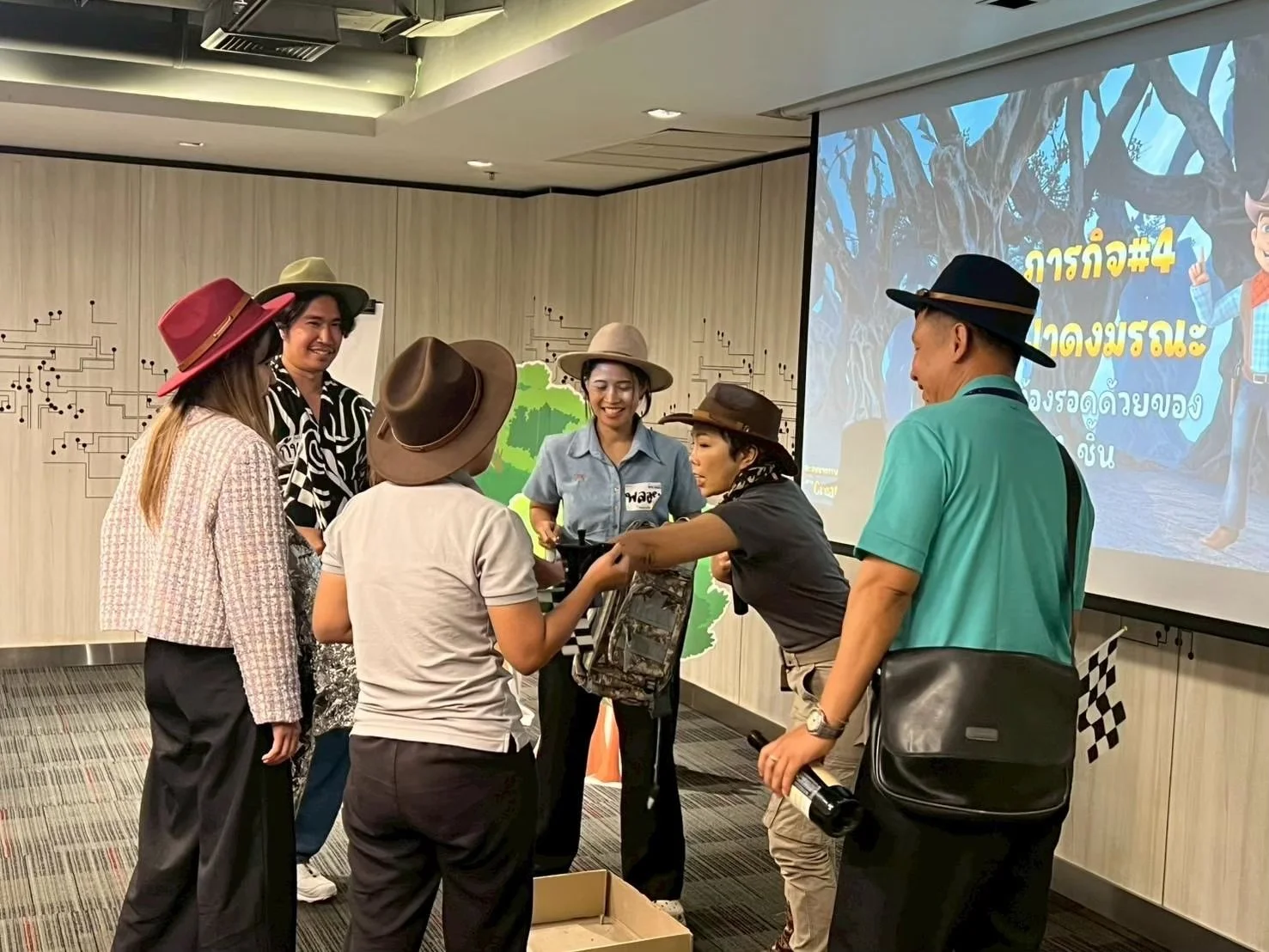 Group of people wearing hats, gathered and engaging in conversation in an indoor setting with a presentation screen in the background.