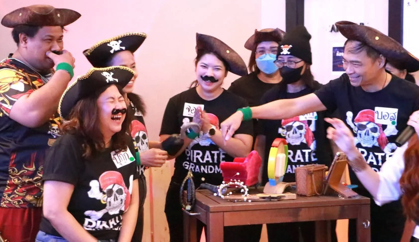 Group of people wearing pirate-themed shirts and hats, laughing and joking around during a party or celebration, with some wearing masks and fake mustaches.