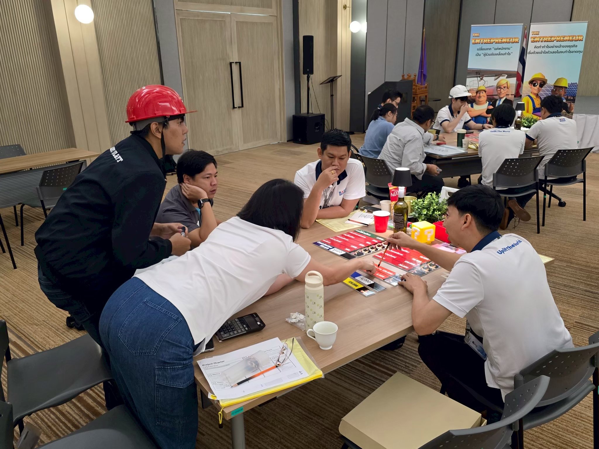 People playing a tabletop game, with some participants wearing white shirts, and a woman in a white shirt reaching across the table. A person in a black jacket and red hard hat is standing nearby, observing the game.