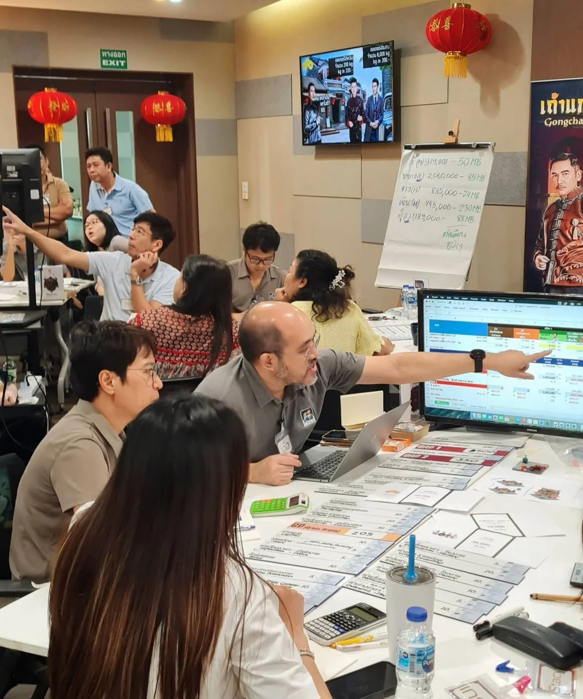 A busy office scene with multiple people working and discussing around a table covered with papers, charts, and laptops. A man in a gray shirt is pointing at a large computer monitor displaying a spreadsheet. Red lanterns and a whiteboard with writing are visible in the background, along with a television screen and a poster.