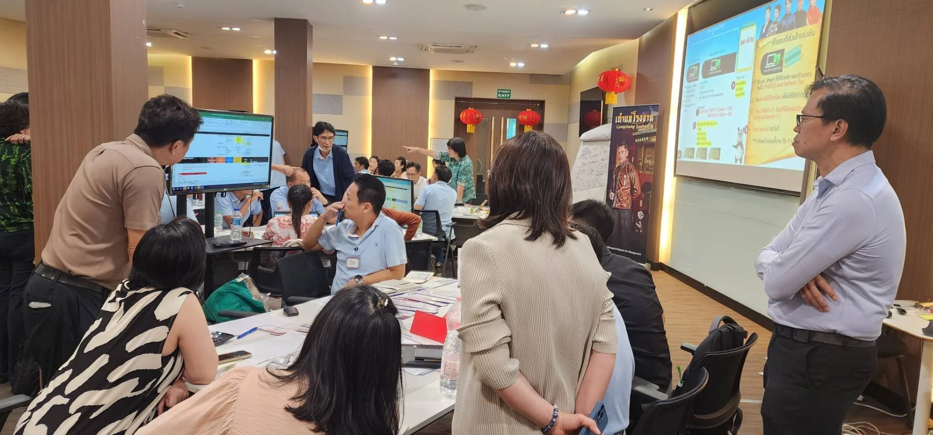 A busy indoor conference or workshop with multiple participants engaged in discussions and watching presentations, featuring large screens and red Chinese lantern decorations.