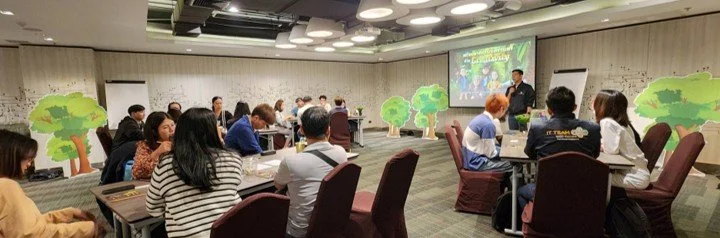 People attending a conference or seminar in a modern room with a speaker presenting at the front, decorated with tree cutouts.