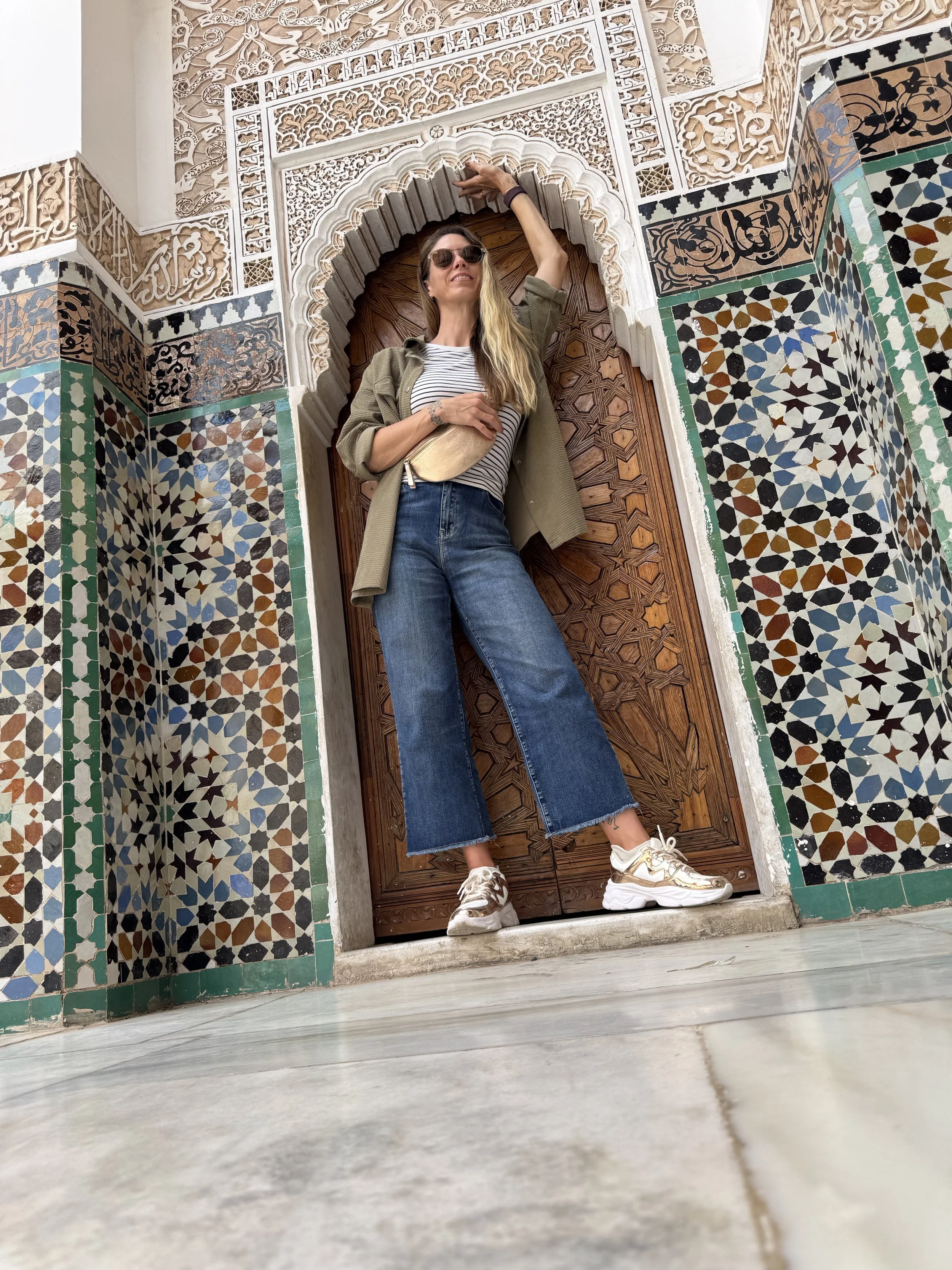 A woman leaning against a decorative wooden door frame with ornate tilework, wearing sunglasses, a striped shirt, an olive green jacket, and denim jeans, in a setting with intricate architectural details.