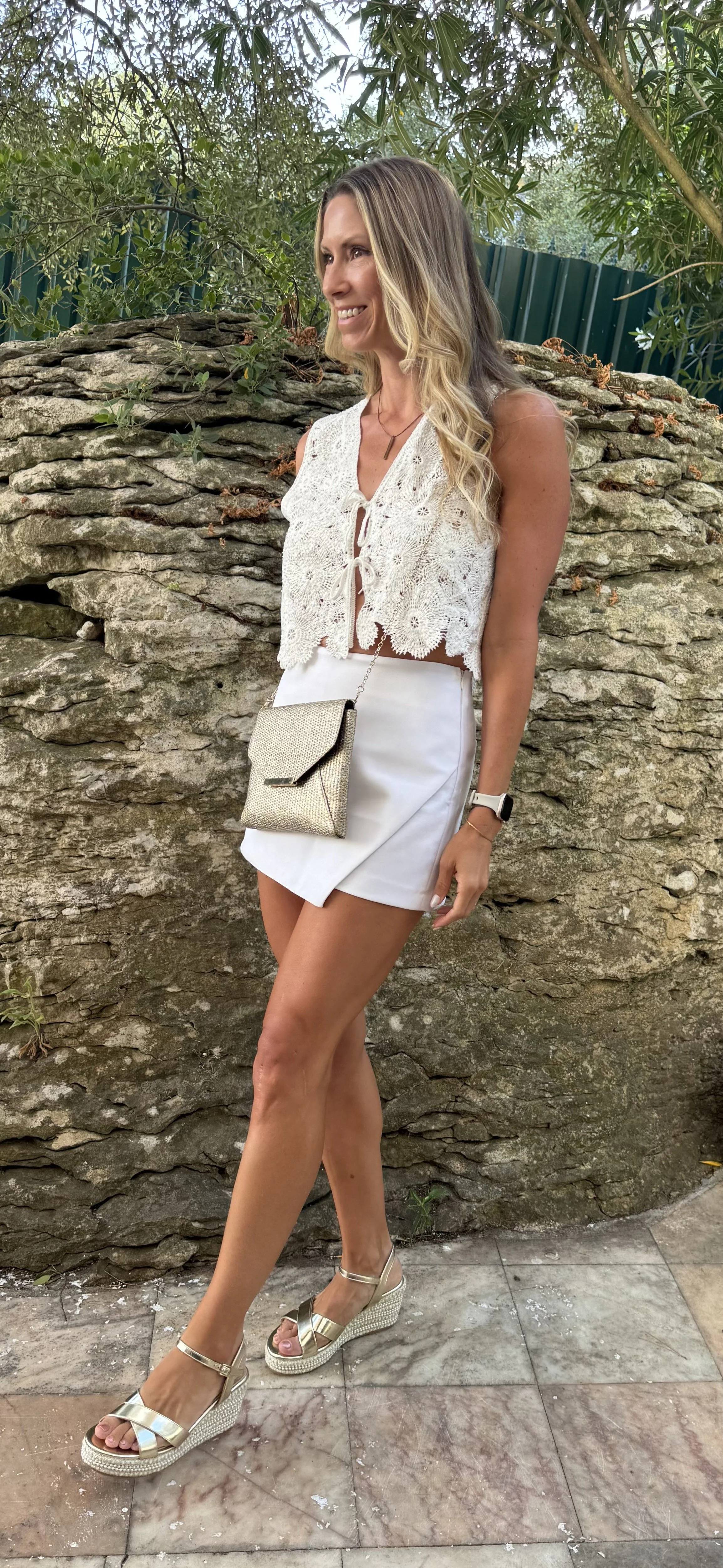 A woman with long wavy blonde hair wearing a white lace sleeveless top, white skirt, beige high-heeled sandals, a small beige purse, a white watch, and a gold bracelet, standing outdoors near a stone wall with greenery in the background.