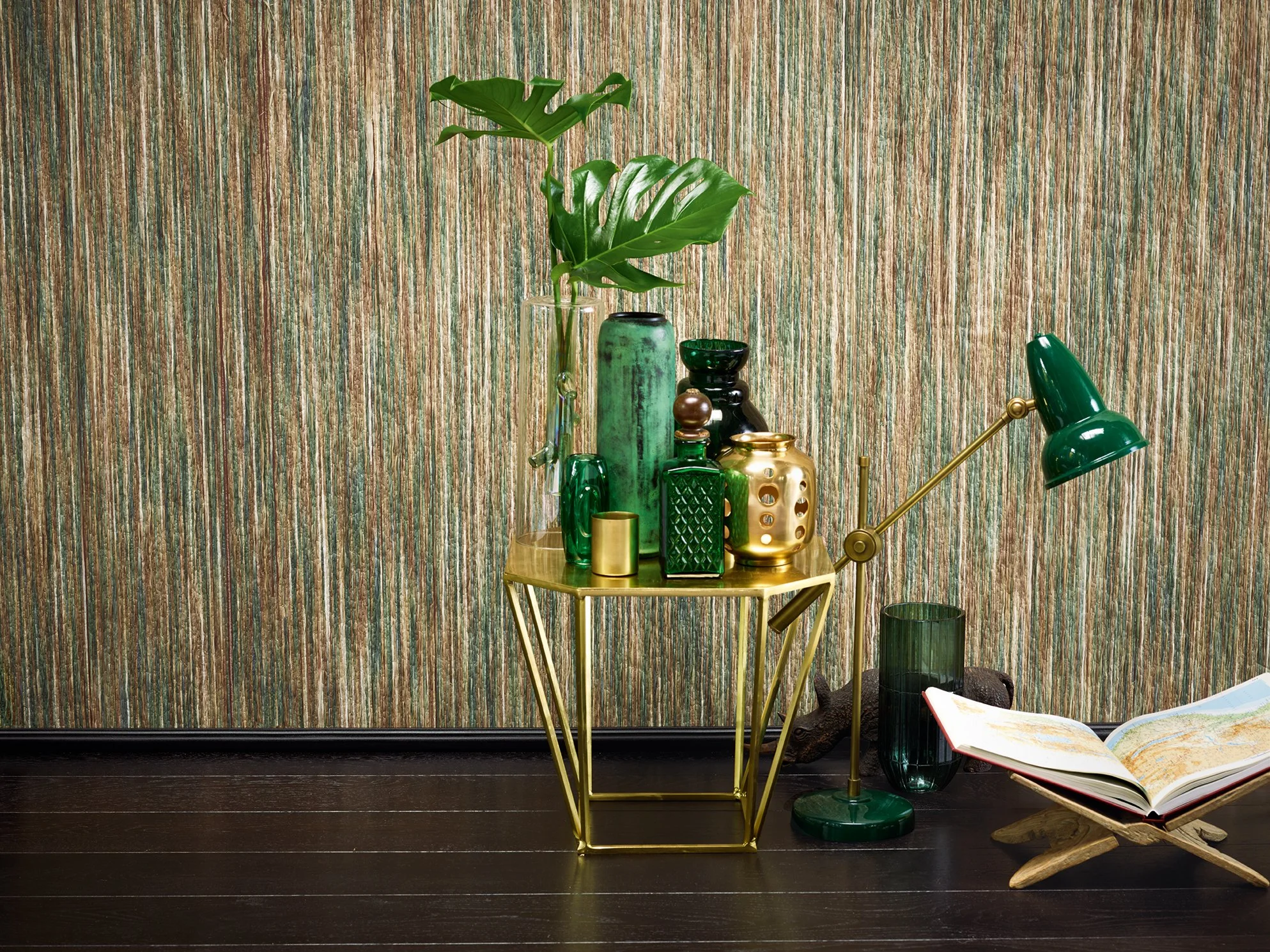 Dekorative Vasen und Kerzen auf einem goldenen Tisch mit einer grünen Pflanzen und einer Leseliege auf dunklem Boden vor einer gemusterten Wand.