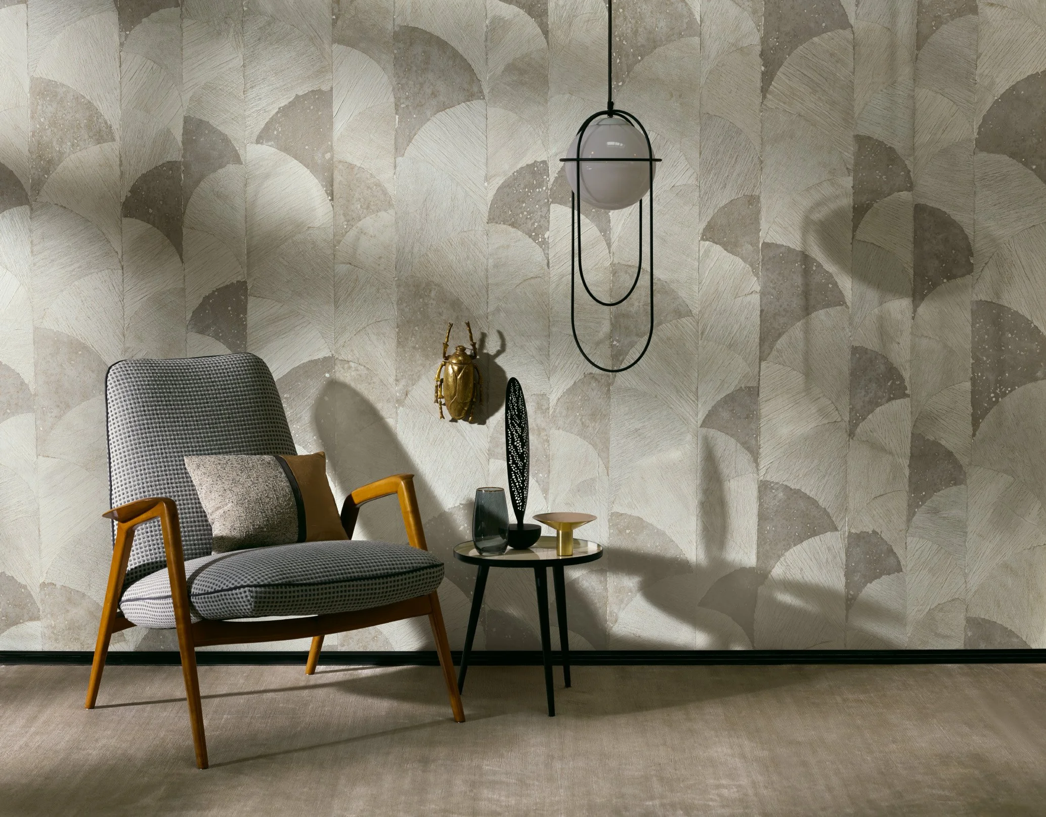 Ein gemütliches Wohnzimmer mit einem modernen Sessel, einer kleinen schwarzen Beistelltisch mit Dekorationsgegenständen, einer goldfarbenen Skulptur, einer schwarzen Lampe und einer goldfarbenen Marienkäferfigur an der Wand. Die Tapete ist in hellen, grafischen Mustern gestaltet.