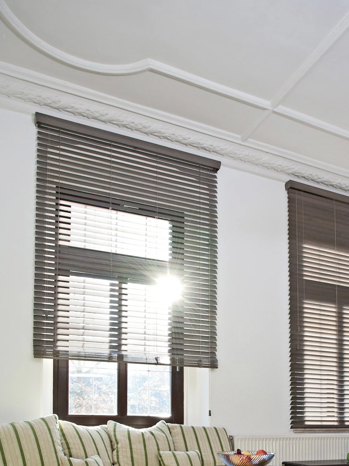 Ein Wohnzimmer mit Fenstern, die mit braunen Jalousien bedeckt sind, und ein Sofa mit grünen und weißen Streifen. Die Sonne scheint durch das Fenster.