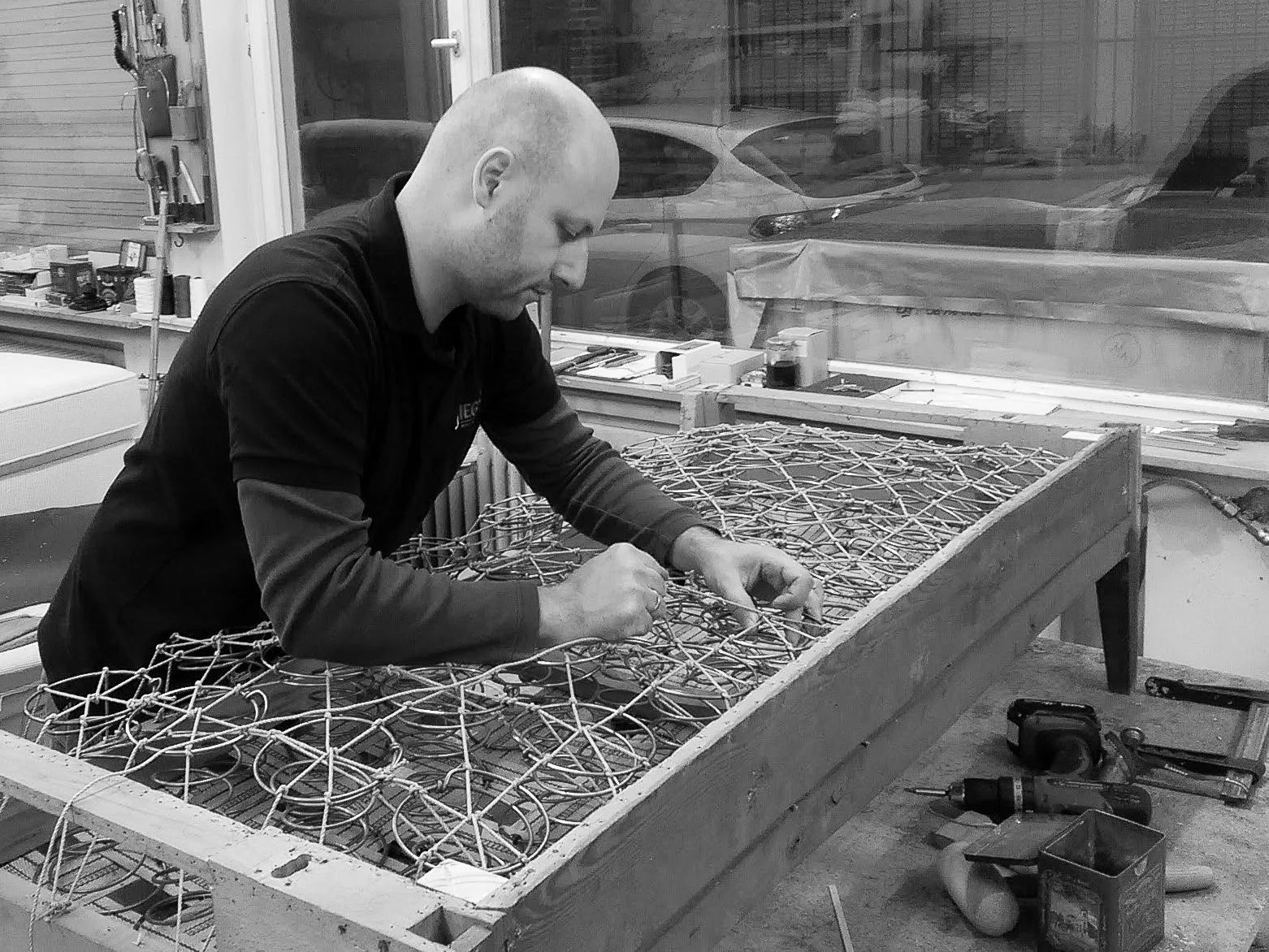 Ein Mann arbeitet in einer Werkstatt an einem Drahtgestell auf einem Tisch, umgeben von Werkzeugen und Materialien.