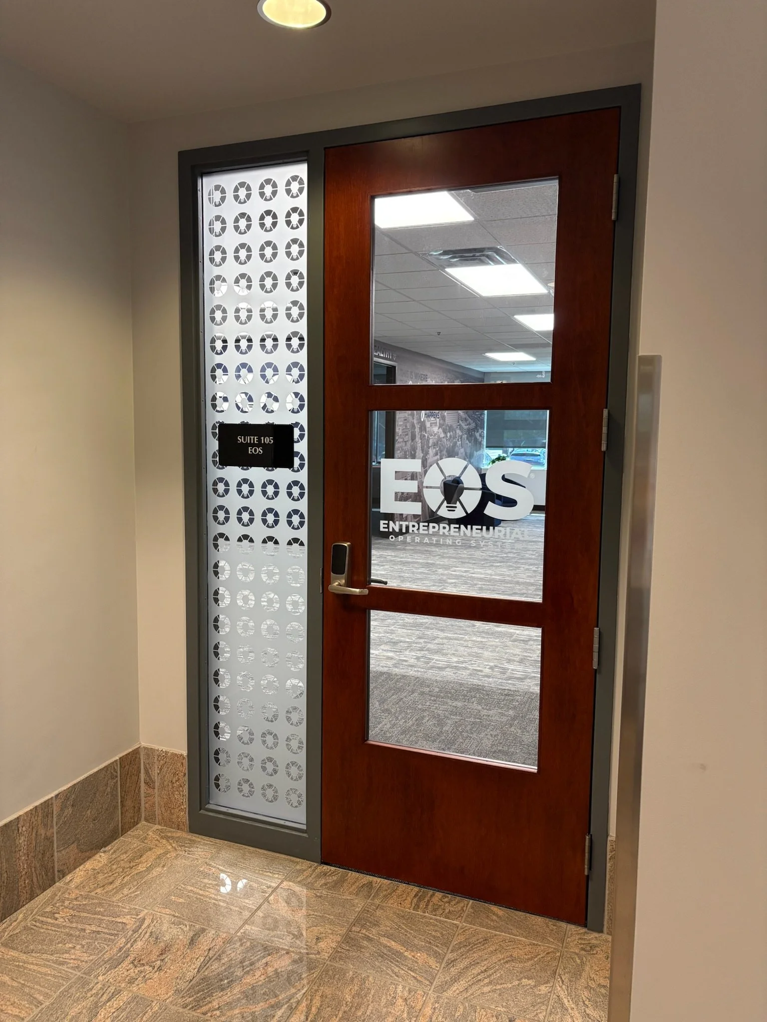 EOS sessions space in Ann Arbor, MI with Interior office door with a glass panel and logo reading EOS ENTREPRENEURIAL OPERATING SYSTEM, next to a decorative metal panel with circular cutouts. A sign indicates Suite 105.