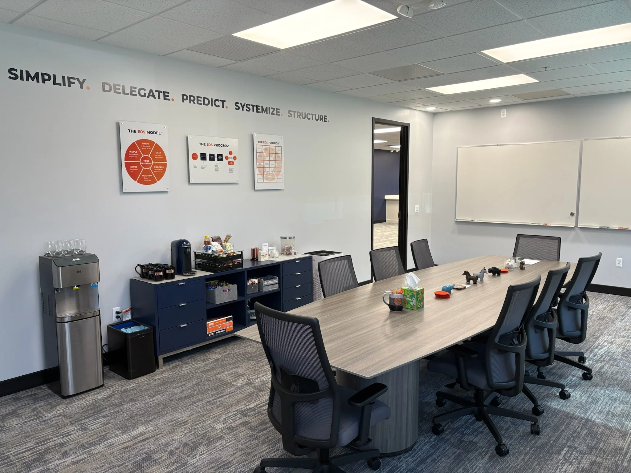 Empty conference room with a long table and chairs, whiteboard, coffee station, water fountain, and wall posters with business process diagrams.