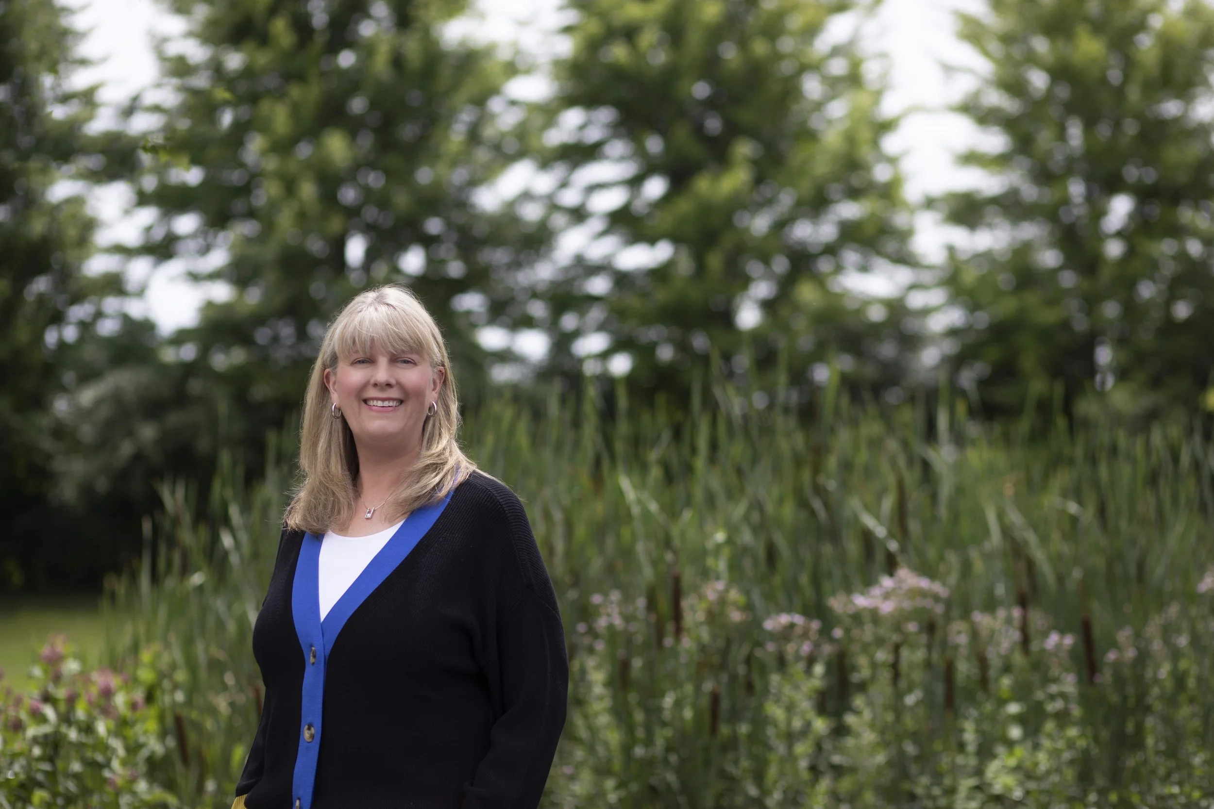 A smiling woman with blonde hair standing outdoors in front of green trees and tall grass or reeds.
