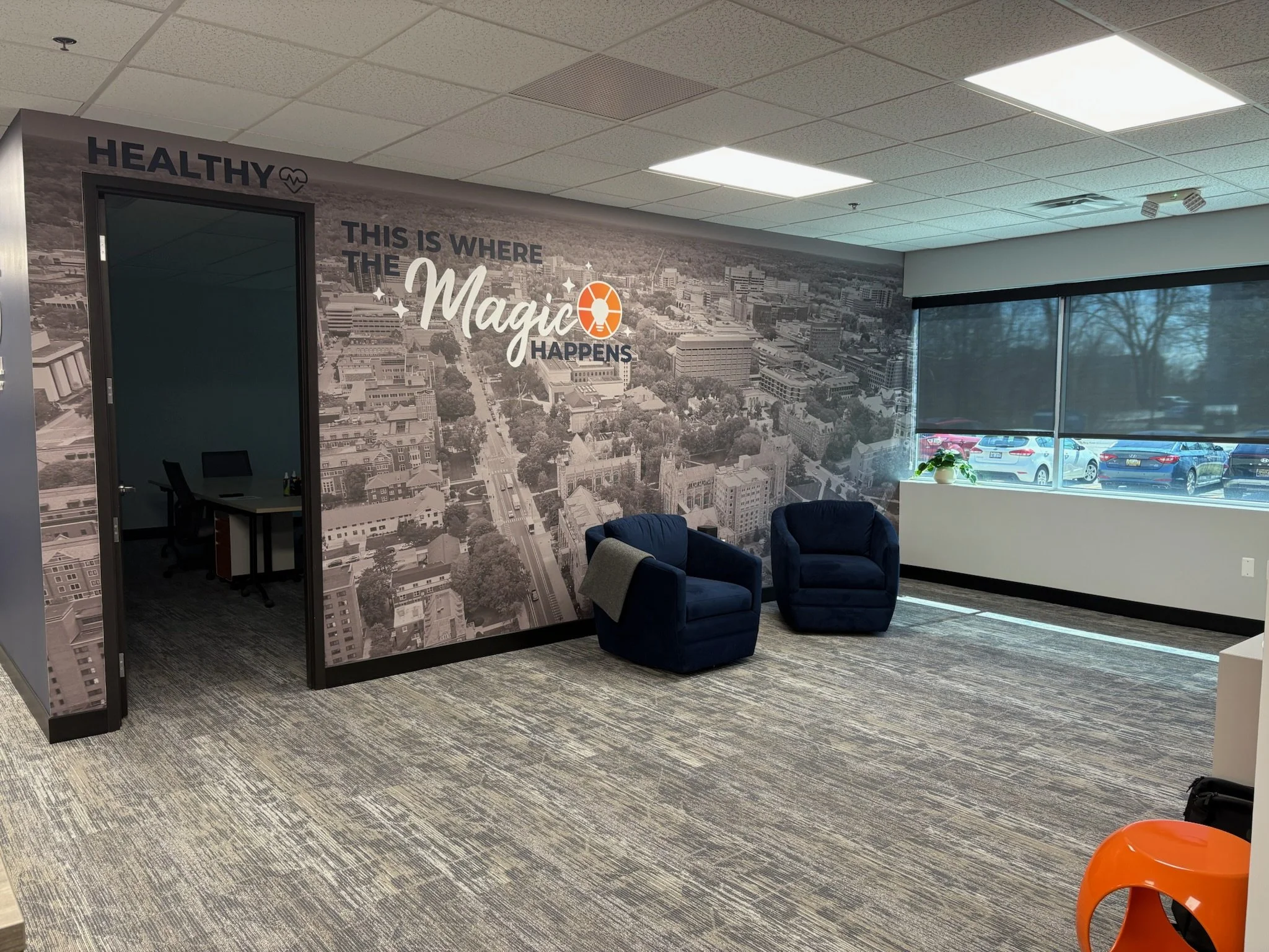 Office lounge area with two blue armchairs and a large window with closed blinds. A black-and-white cityscape mural on the wall features the quote "This is where the magic happens" with the word "magic" highlighted and a small orange circle with a wh