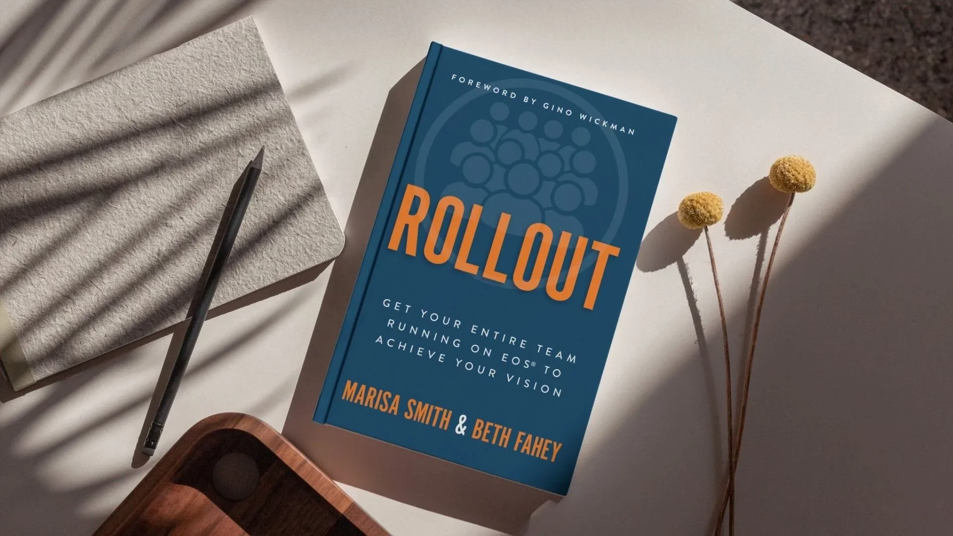 A book titled 'Rollout' by Marisa Smith and Beth Fahey is on a white surface. The cover is blue with orange and white text. To the left of the book is a textured beige notepad with a black pen resting on it. To the right of the book are two yellow billy balls flowers casting shadows. Part of a wooden object is visible in the bottom left corner.