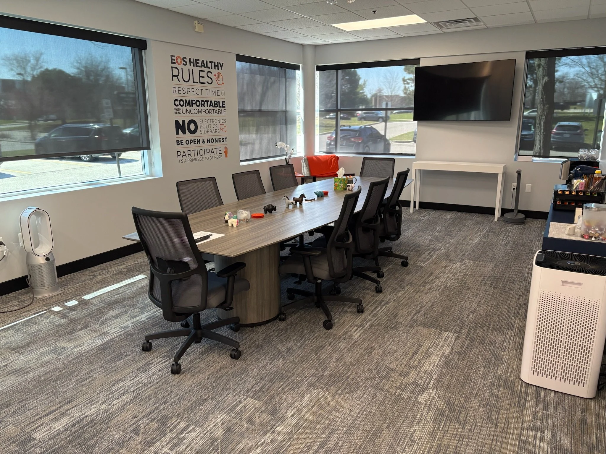 Conference room with a long table, eight black office chairs, and several small toys and items on the table. There are large windows with black blinds, a wall poster with health rules, a large flat-screen TV on the wall, a white rectangular table, a 