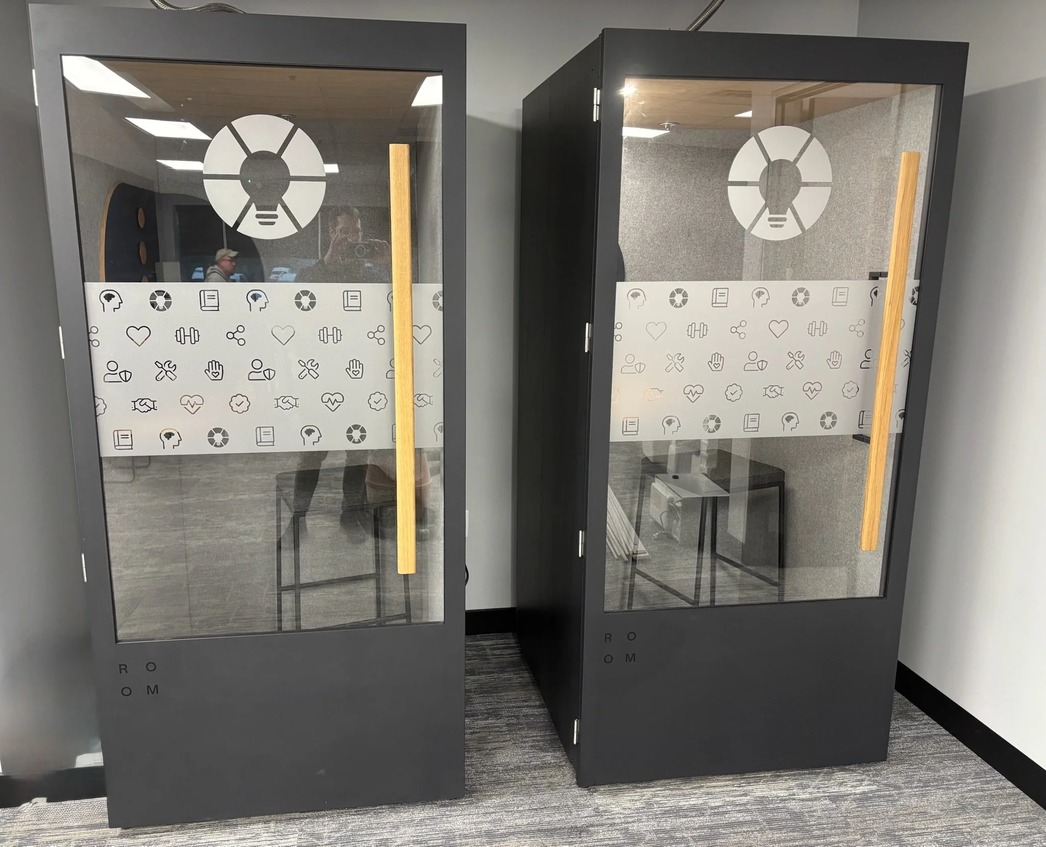 EOS sessions space in Ann Arbor, MI, with Two black and glass phone booths with wooden door handles, decorated with health and wellness icons, in a modern office setting.