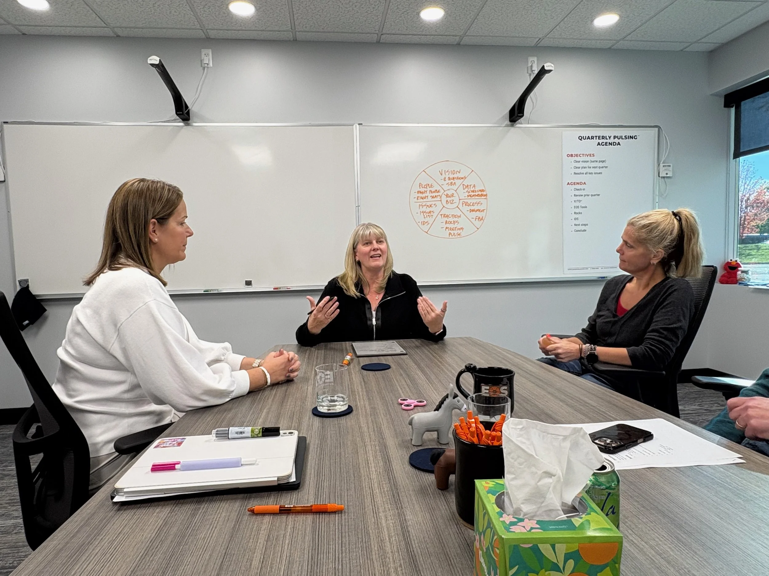 Three women in a meeting room having a discussion at a large table. The woman in the middle appears to be speaking, while the other two listen attentively. The table has notebooks, pens, a glass of water, tissues, and office supplies. A whiteboard with notes and a schedule is behind them, and a window shows trees outside.