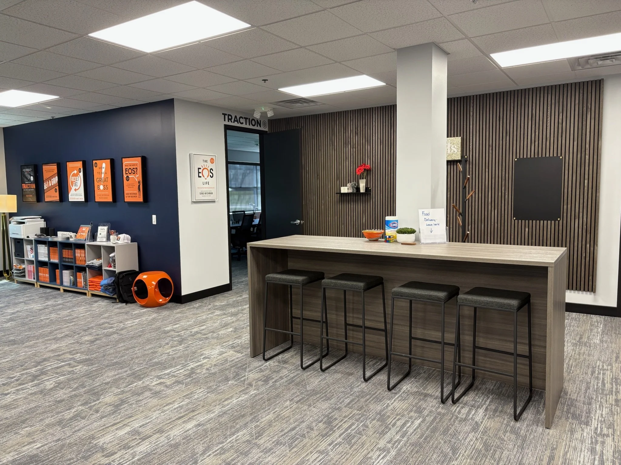 Office reception area with a high table, four barstools, a dark wood-paneled wall with a shelf, a flower vase, snacks, and a sign for food delivery. To the left, there is a weathered white shelf with orange books or magazines, a printer, and decorati