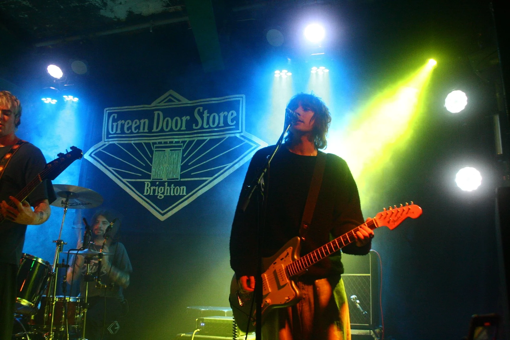 Band performing live on stage at Green Door Store in Brighton, with a bassist, a drummer, and a guitarist under colorful stage lighting