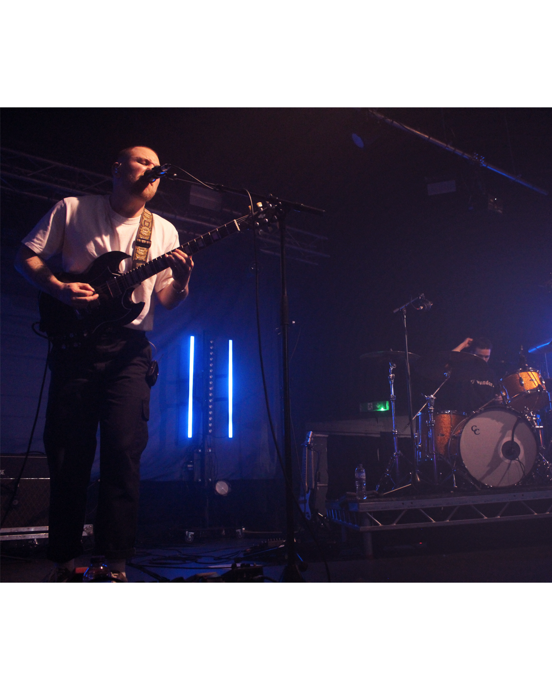 Musician performing on stage with a guitar and singing into a microphone, with a drummer in the background, stage lighting in blue.