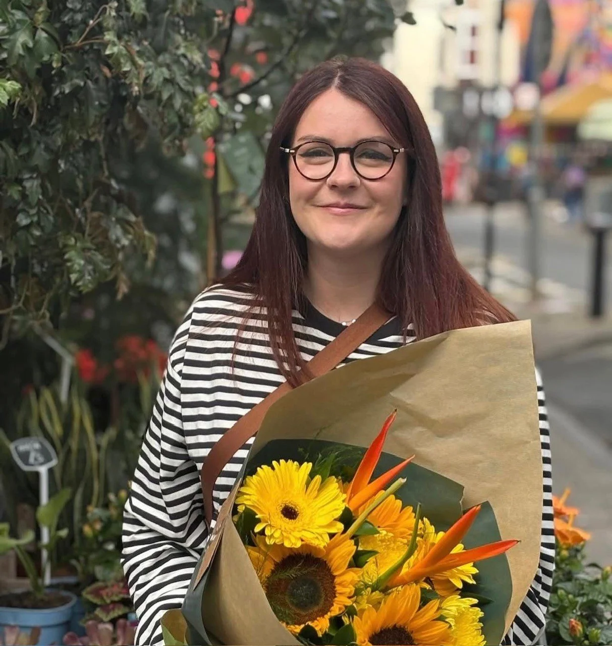 A woman with glasses and long brown hair holding a bouquet of yellow and orange flowers on a city sidewalk.